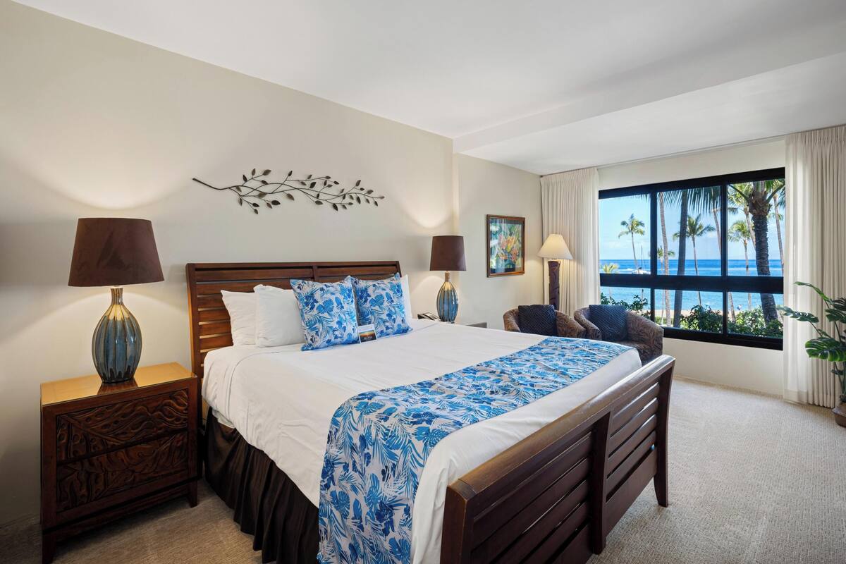 A hotel room with a large bed, two bedside tables, a window with a view of palm trees and the ocean, and a decorative wall piece.
