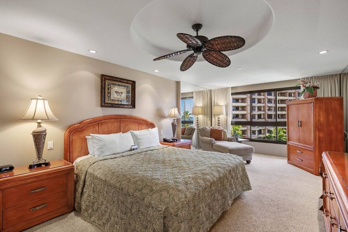 A hotel room with a bed, a nightstand, a desk, a chair, a wardrobe, a ceiling fan, and a window with a view of the hotel.