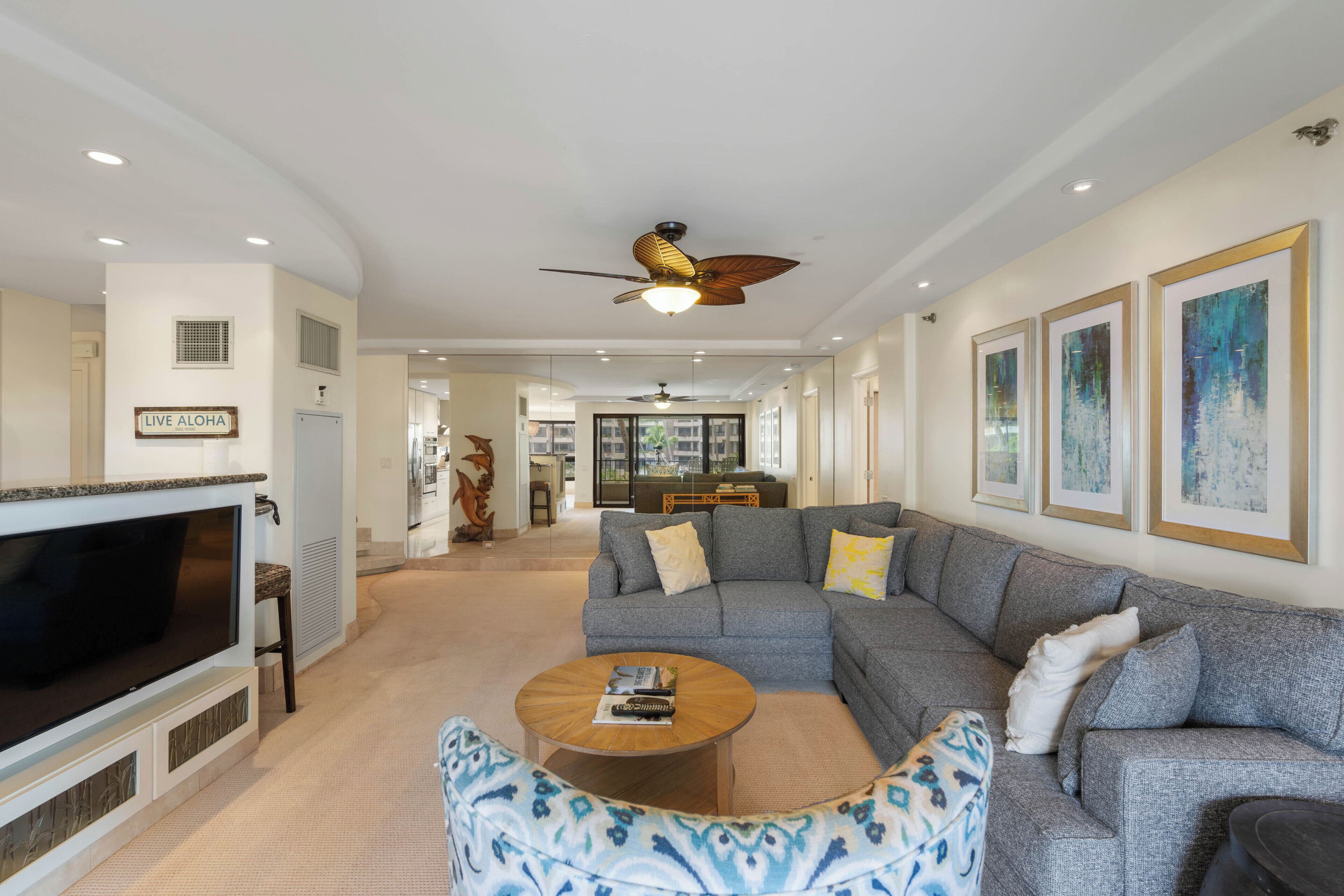 A spacious living room with a large flat-screen TV, a grey sectional sofa, a round coffee table, and a ceiling fan.