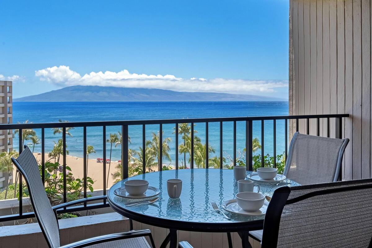 A balcony with a table set for two, overlooking a beach and ocean with a mountain in the distance.