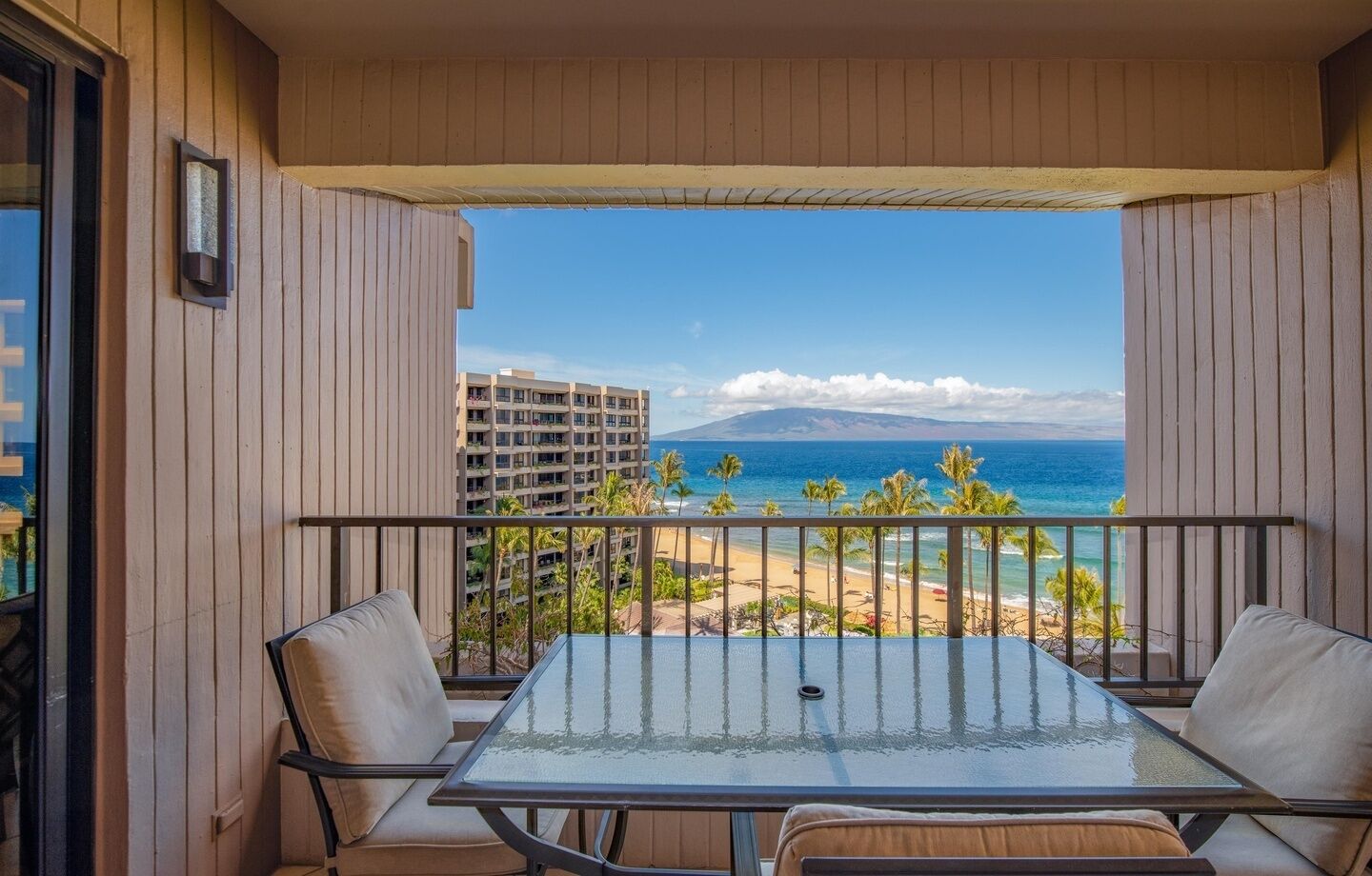 Un balcon avec une table et des chaises en verre, donnant sur une plage et l’océan, avec des bâtiments visibles à l’arrière-plan.