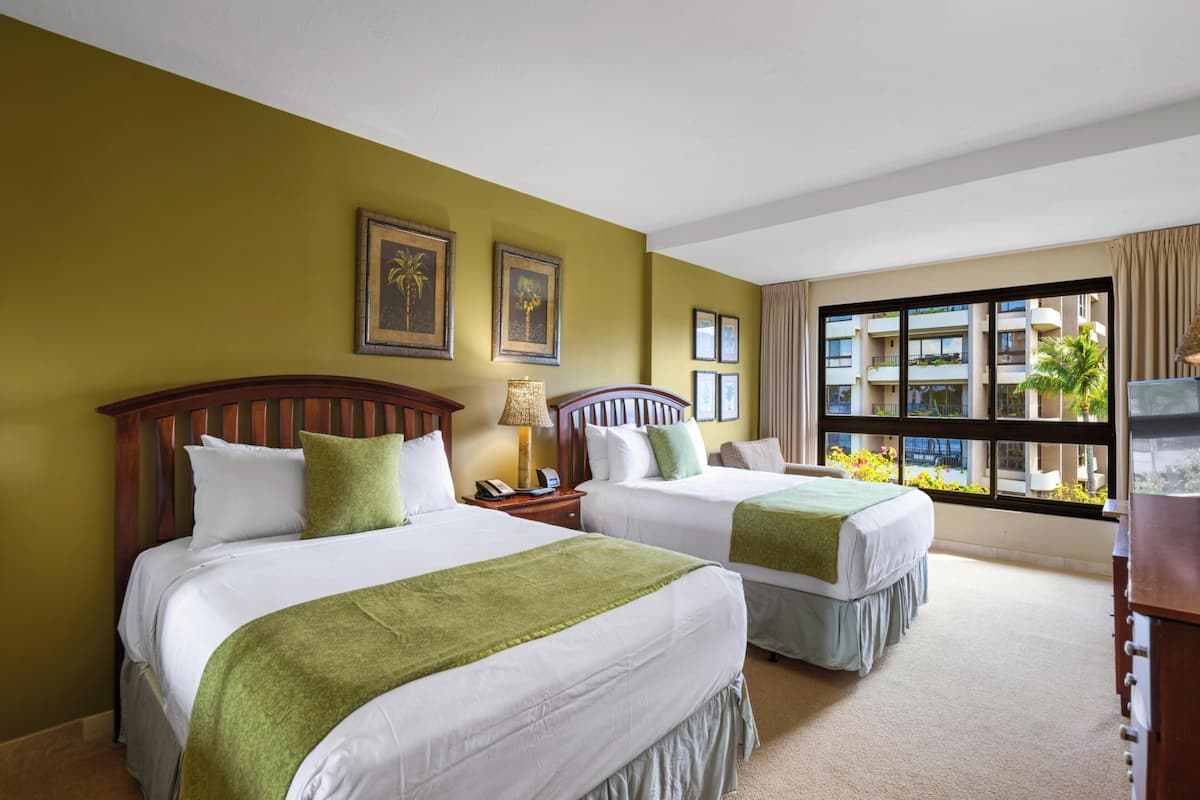 A hotel room with two beds, a large window with a view of the building, and a desk with a lamp.