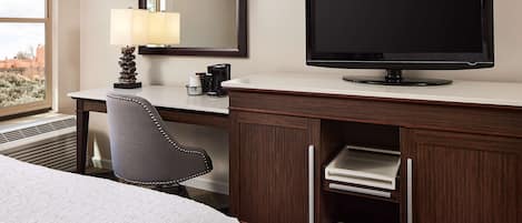 A hotel room with a bed, a desk with a computer monitor, a lamp, a chair, and a large mirror.