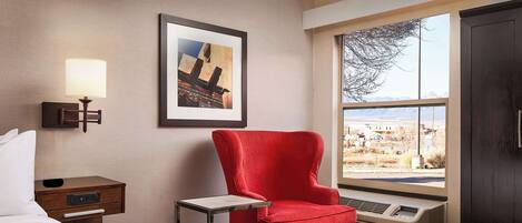 A hotel room with a bed, a red armchair, a small table, a nightstand, a wall-mounted lamp, a framed picture, a window with a view, and an air conditioning unit.