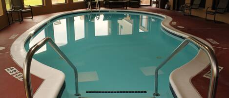 An indoor swimming pool with steps leading into the water, surrounded by chairs and tables.
