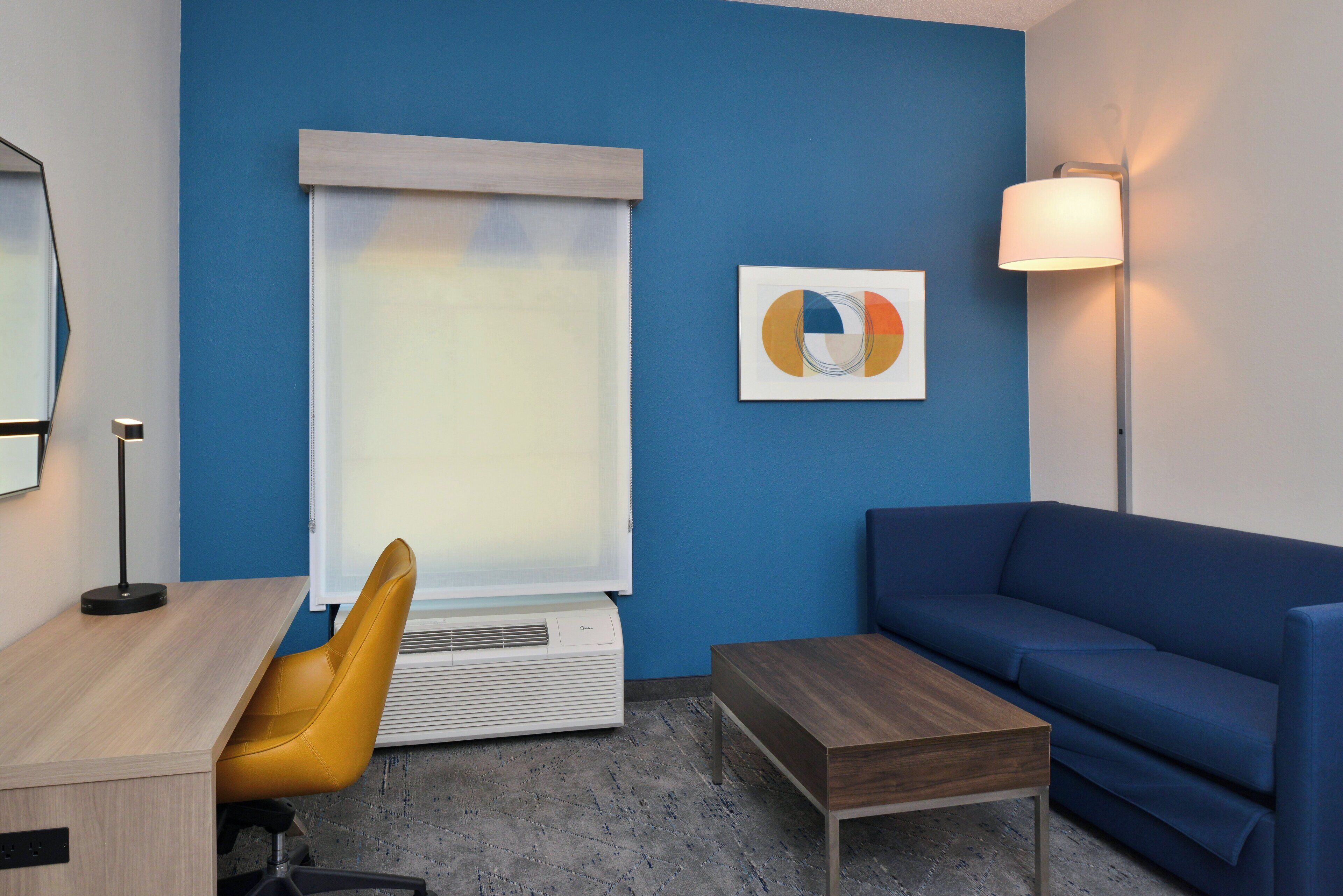 A modern room with a blue wall, a wooden desk, a yellow chair, a blue sofa, a small wooden table, and a floor lamp.