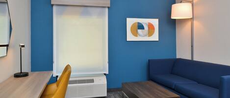 A modern room with a blue wall, a wooden desk, a yellow chair, a blue sofa, a small wooden table, and a floor lamp.