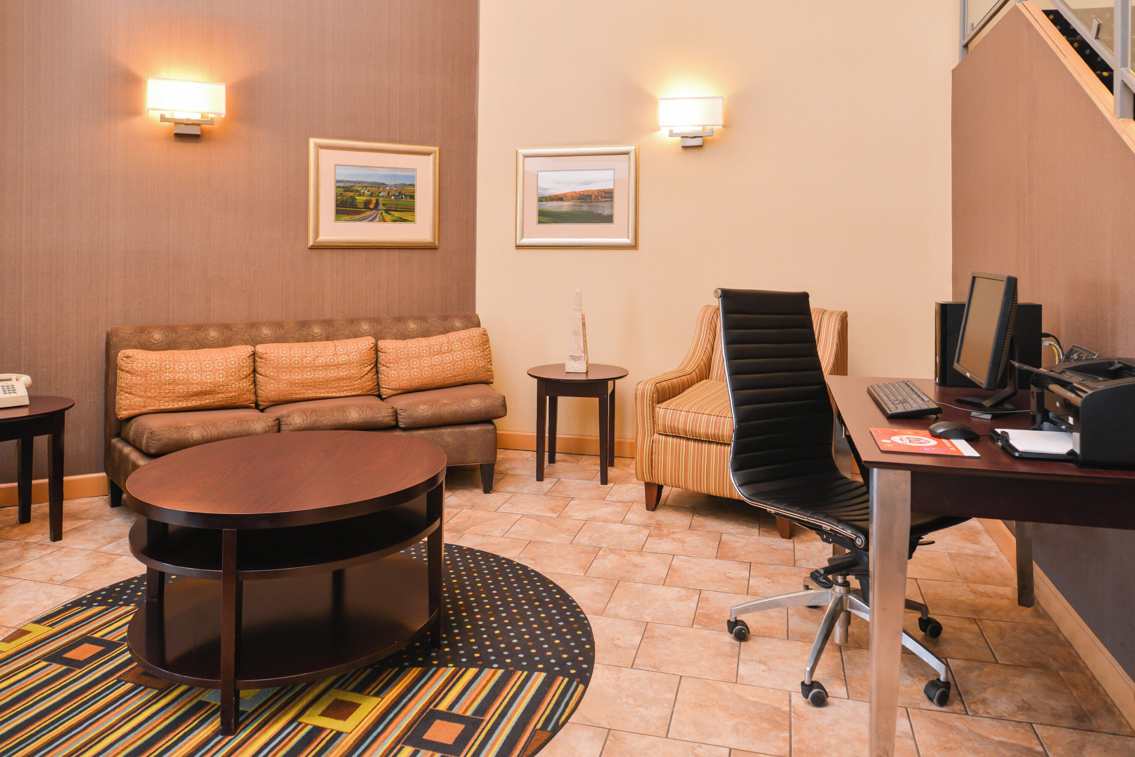 A hotel room with a sofa, a round table, a chair, a desk with a computer, and two wall-mounted lamps.