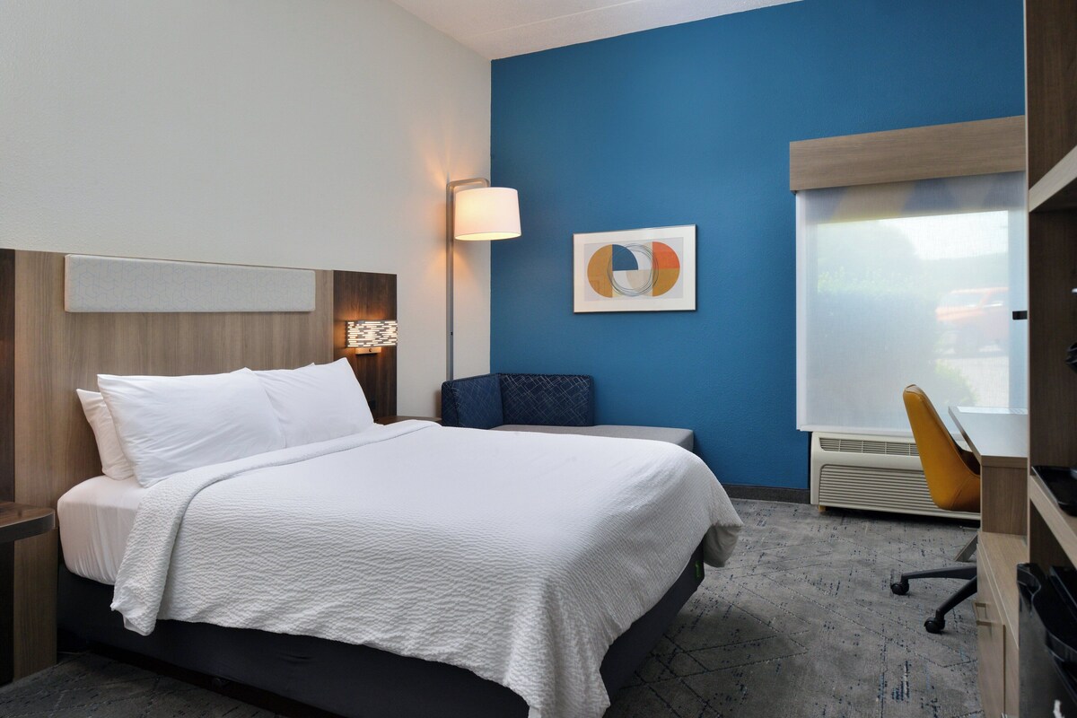 A hotel room with a bed, a desk, a chair, a wall-mounted lamp, and a window with blinds.