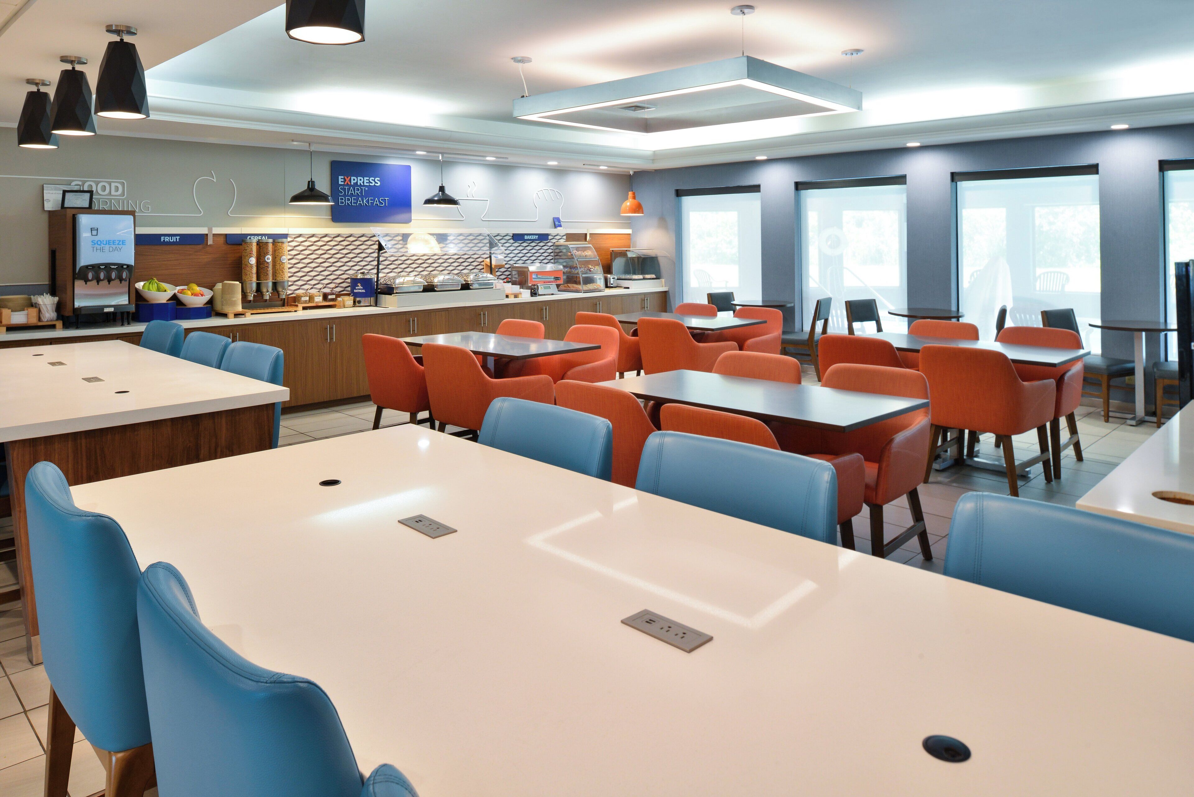 Un área de comedor moderna con mesas blancas y sillas azules y anaranjadas, una estación de café y ventanas amplias.