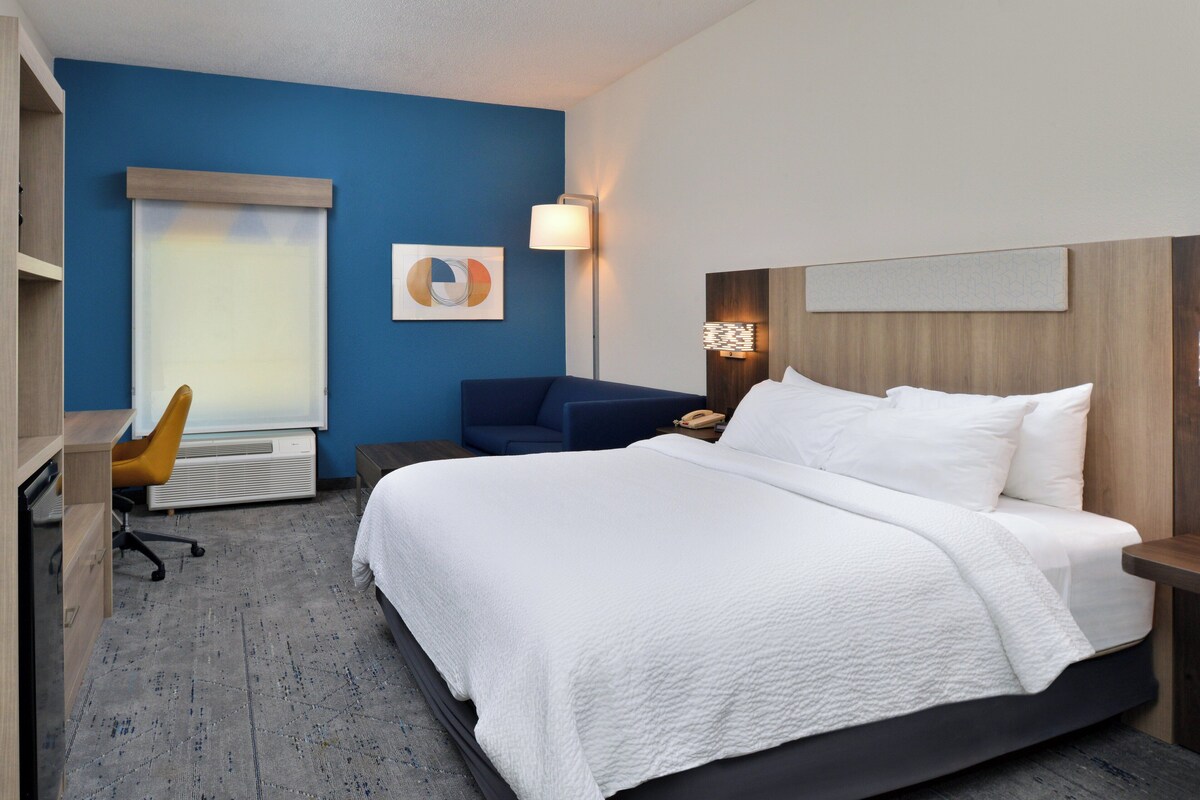 A hotel room with a large bed, a desk with a chair, a sofa, and a wall-mounted lamp.