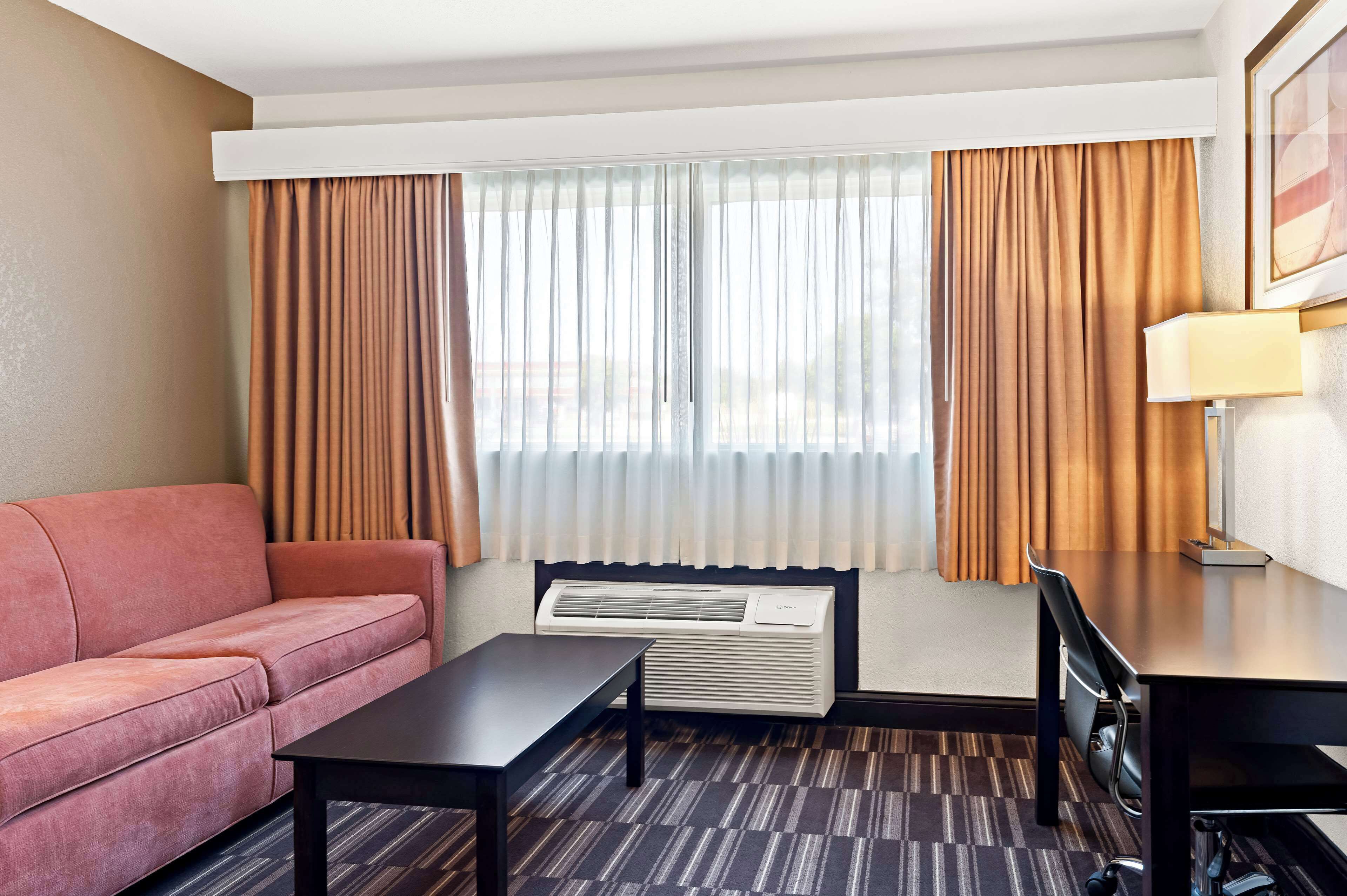 A hotel room with a sofa, a table, a desk, a lamp, and an air conditioning unit.