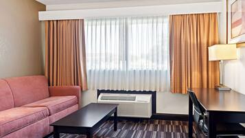 A hotel room with a sofa, a table, a desk, a lamp, and an air conditioning unit.