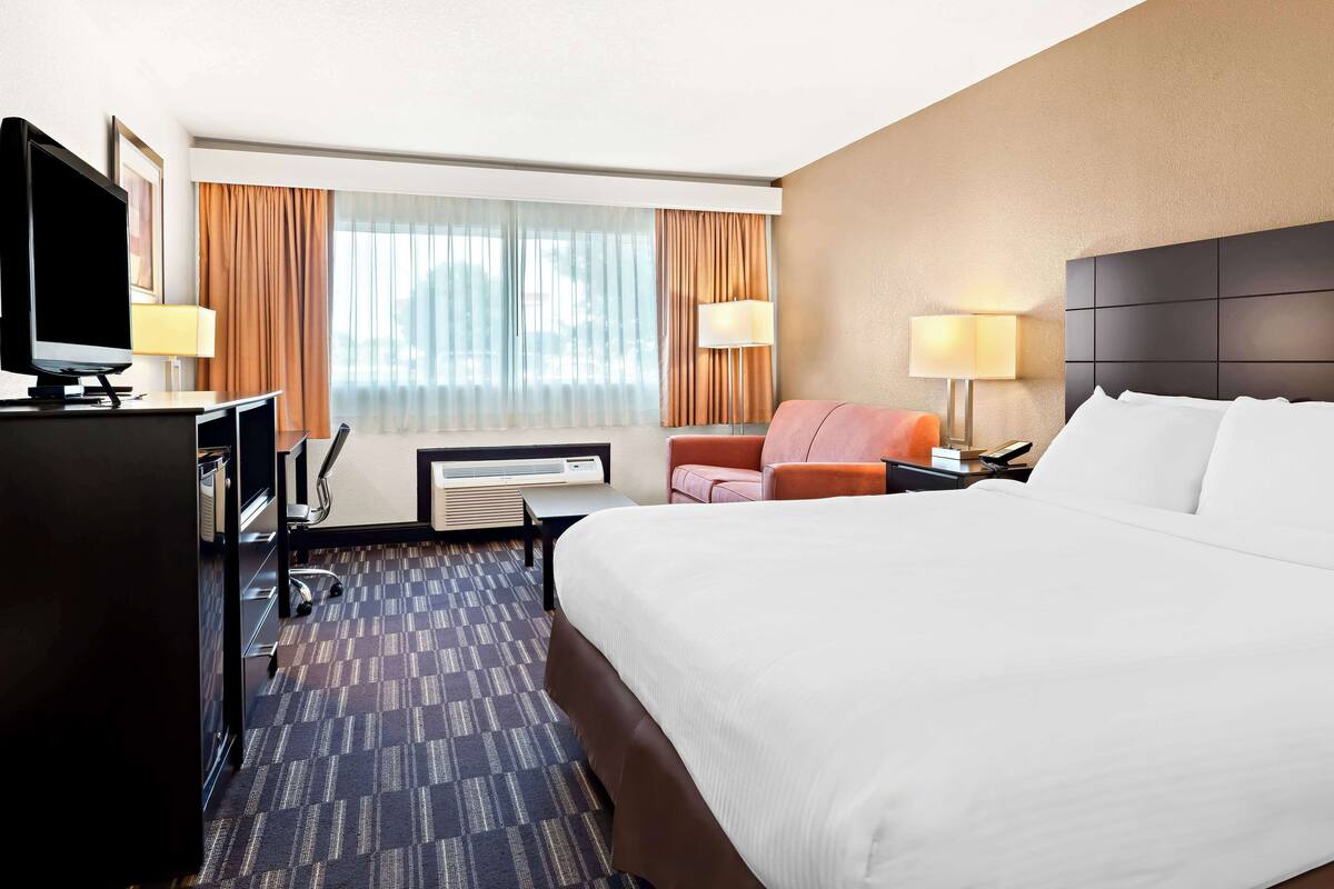 A hotel room with a large bed, a desk with a computer, a TV, two chairs, and a window with curtains.