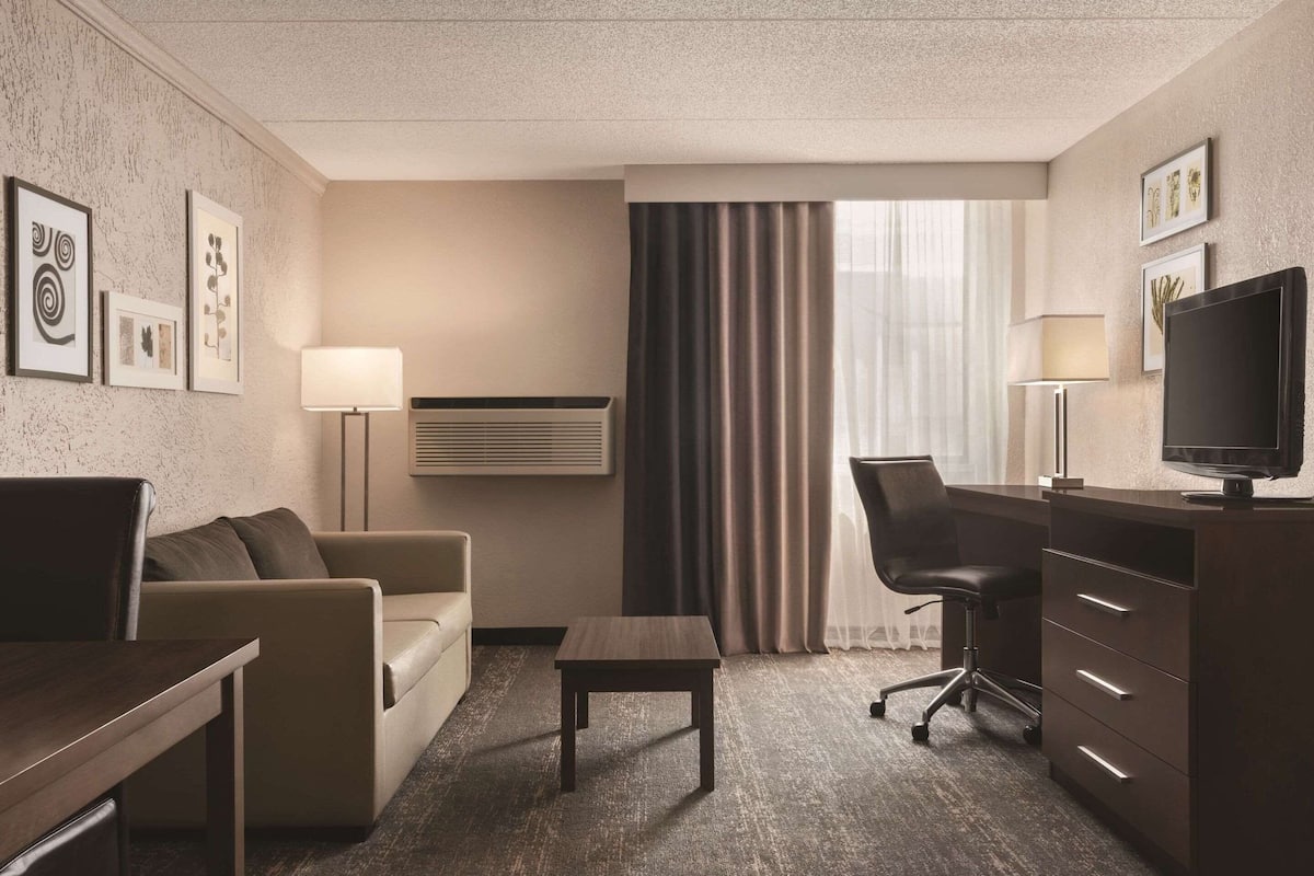 Quarto de hotel com sofá, mesa pequena, escrivaninha com cadeira, televisão e janela com cortinas.