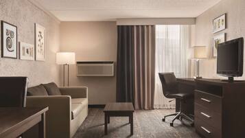 A hotel room with a sofa, a small table, a desk with a chair, a TV, and a window with curtains.