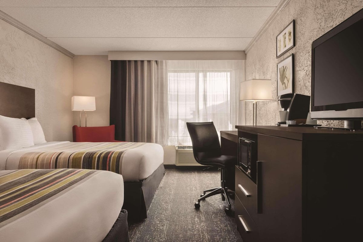 Quarto de hotel com duas camas, uma escrivaninha, uma cadeira, uma televisão e uma janela com cortinas.