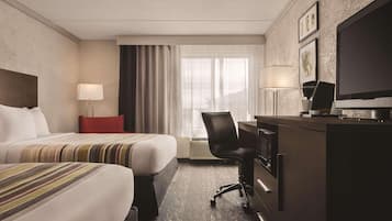 Ein Hotelzimmer mit zwei Betten, einem Schreibtisch, einem Stuhl, einem Fernseher und einem Vorhangfenster.