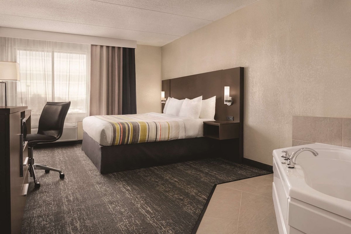 Quarto de hotel com cama, escrivaninha, cadeira, luminária de mesa, mesa de cabeceira, abajur, janela com cortinas e banheira.