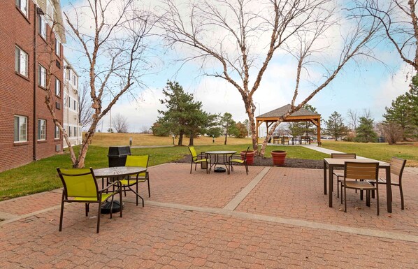 An outdoor patio area with brick flooring, green and yellow chairs, and a wooden gazebo.