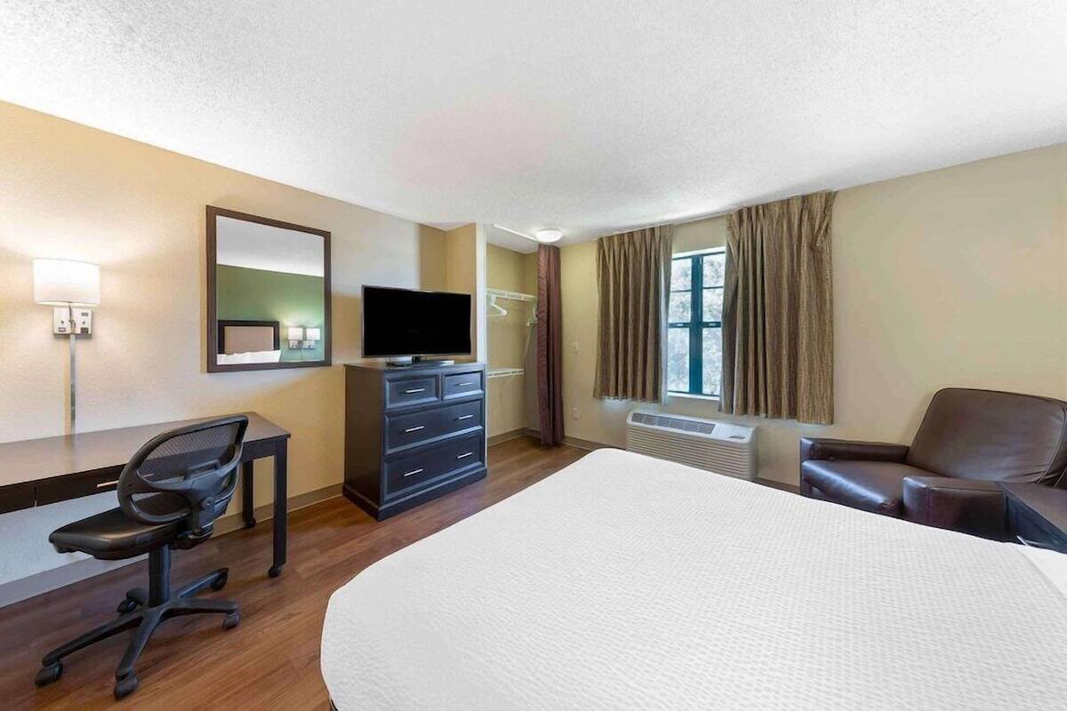 A hotel room with a large bed, a desk with a chair, a television, a dresser, and a window with curtains.