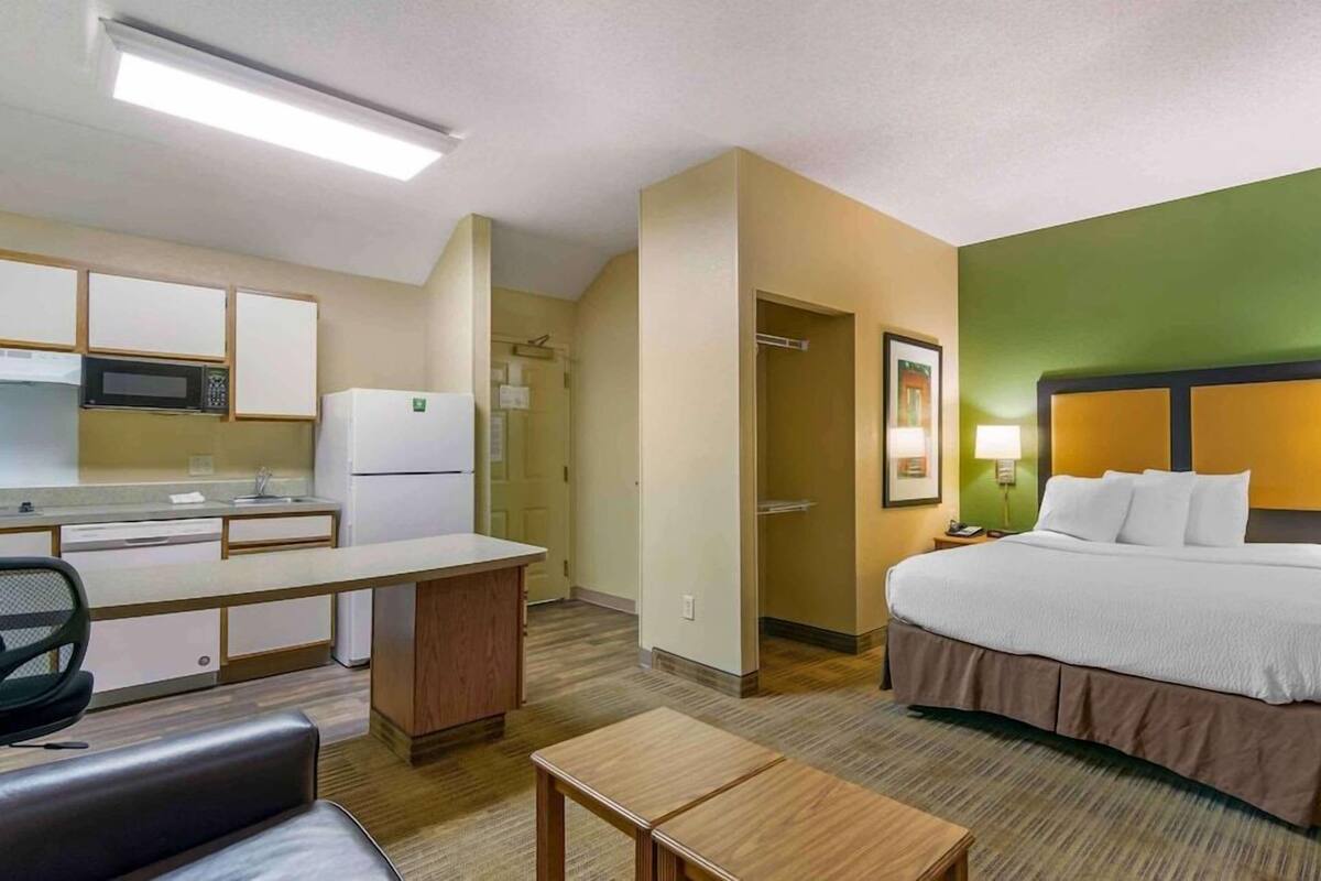 A hotel room with a bed, a desk, a chair, a microwave, a refrigerator, and a small kitchenette.