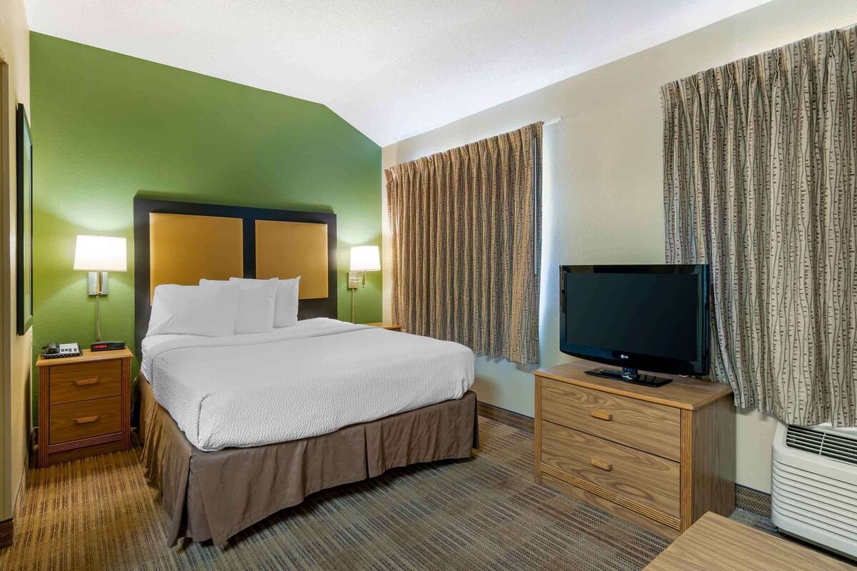 A hotel room with a bed, bedside lamps, a television, a dresser, and a window with curtains.