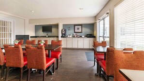 A modern dining area with red upholstered chairs and a wooden table, a kitchen with white cabinets and a flat-screen TV, and large windows with blinds.