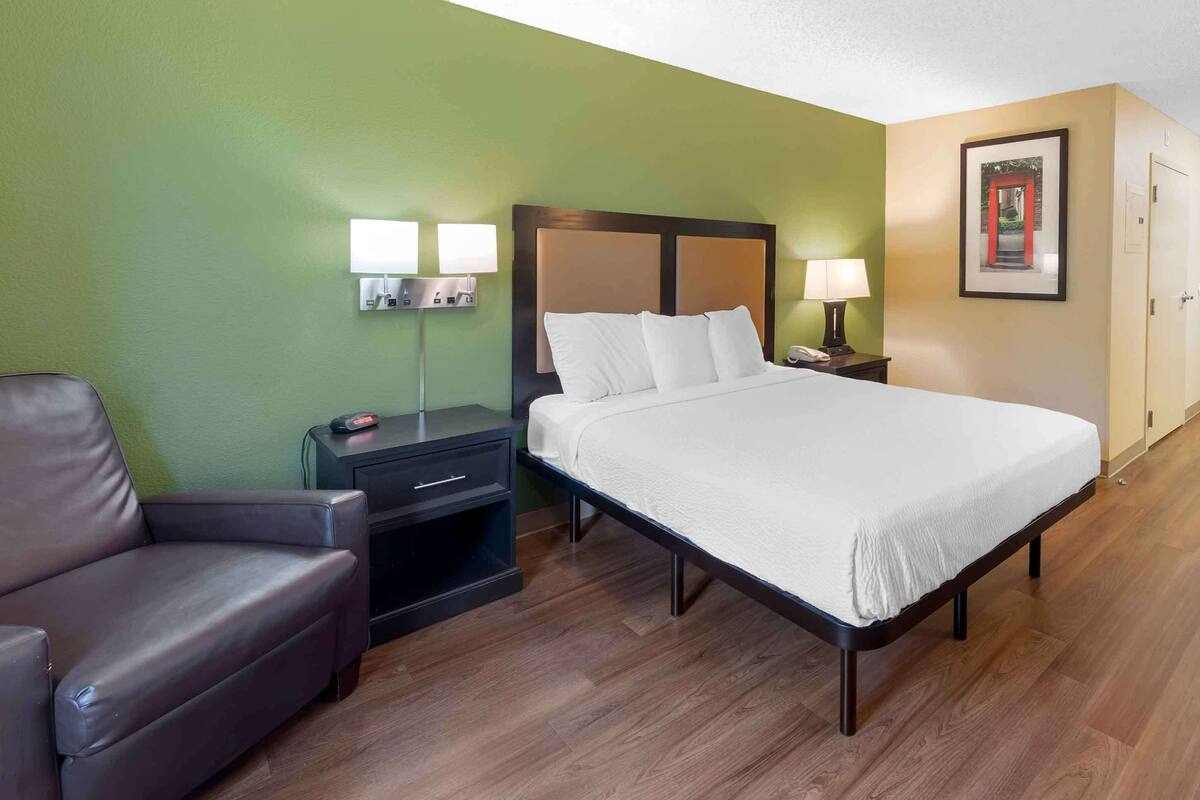 A hotel room with a bed, a nightstand, a lamp, a chair, and a framed picture on the wall.