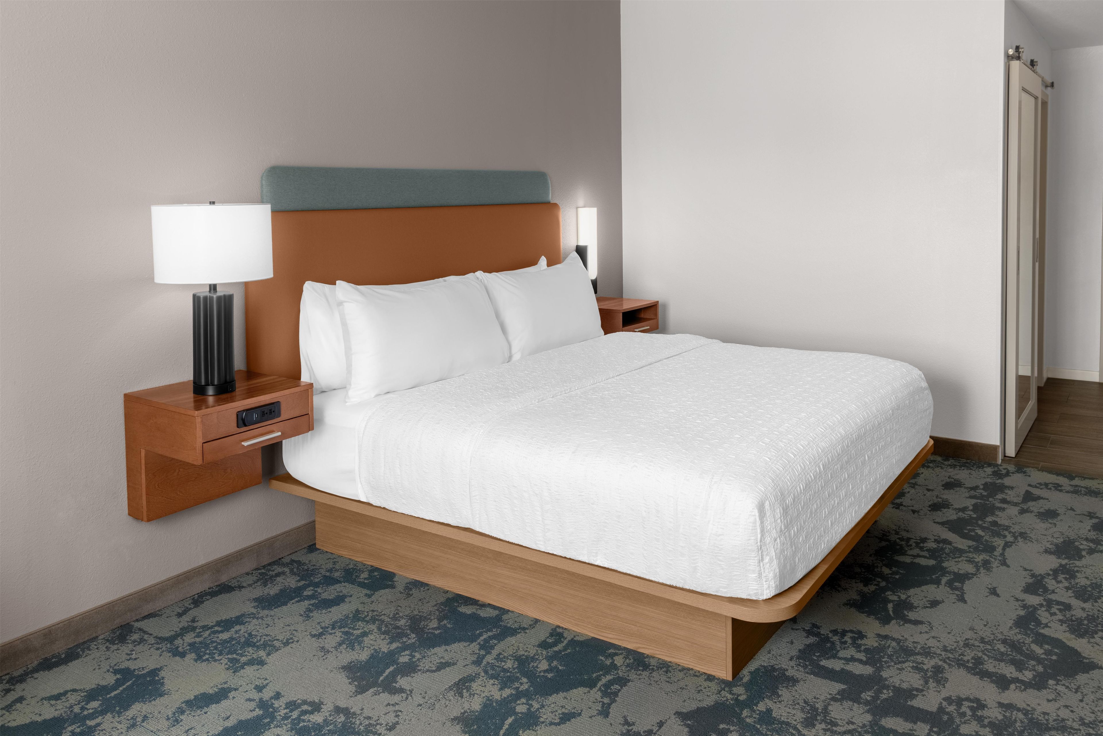 A hotel room with a bed, a nightstand, a lamp, and a wall-mounted light.
