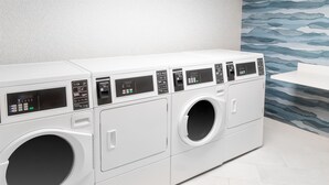 Une rangée de machines à laver à chargement frontal, toutes blanches, dans une buanderie dont le sol est carrelé et un des murs est décoré d’un motif ondulé bleu.