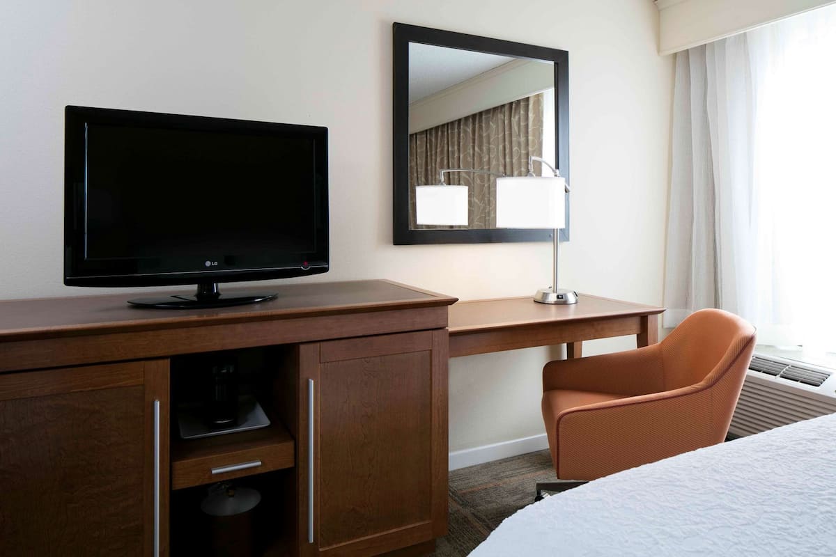 A hotel room with a wooden desk, a television, a mirror, a chair, and a bed.