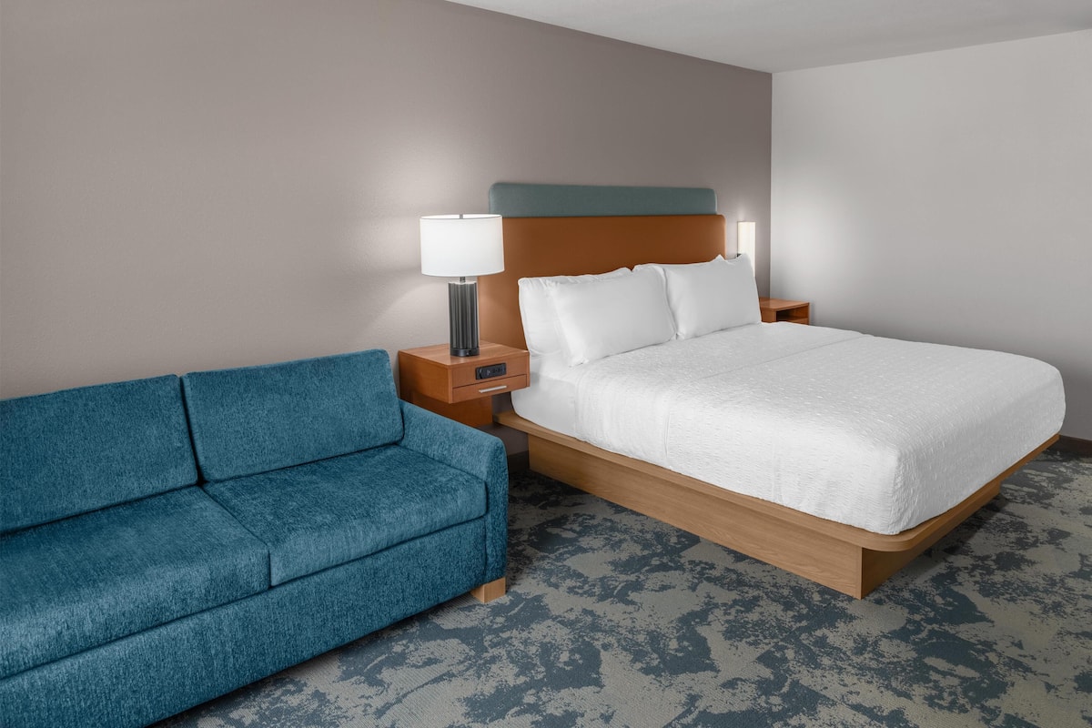 Habitación de hotel con una cama, un sofá azul, una mesita de noche con lámpara y un cabecero de madera.