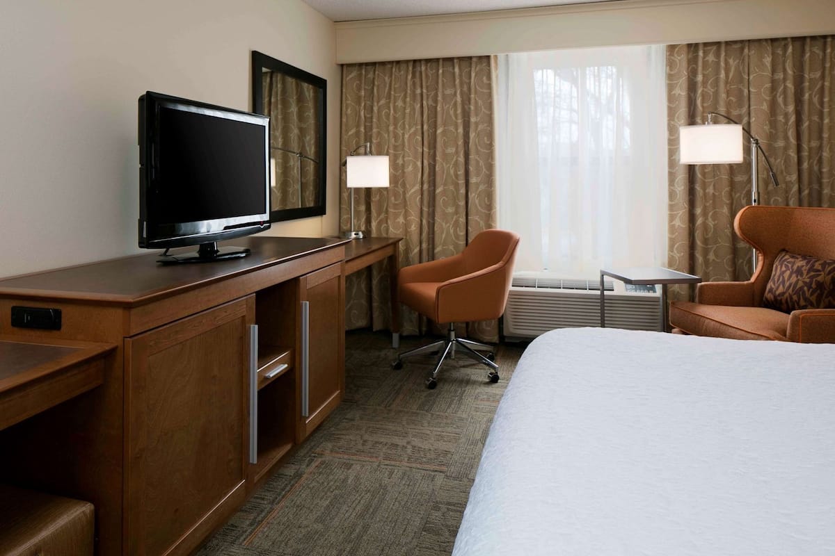 A hotel room with a bed, a desk with a television, a chair, a small table, and a window with curtains.