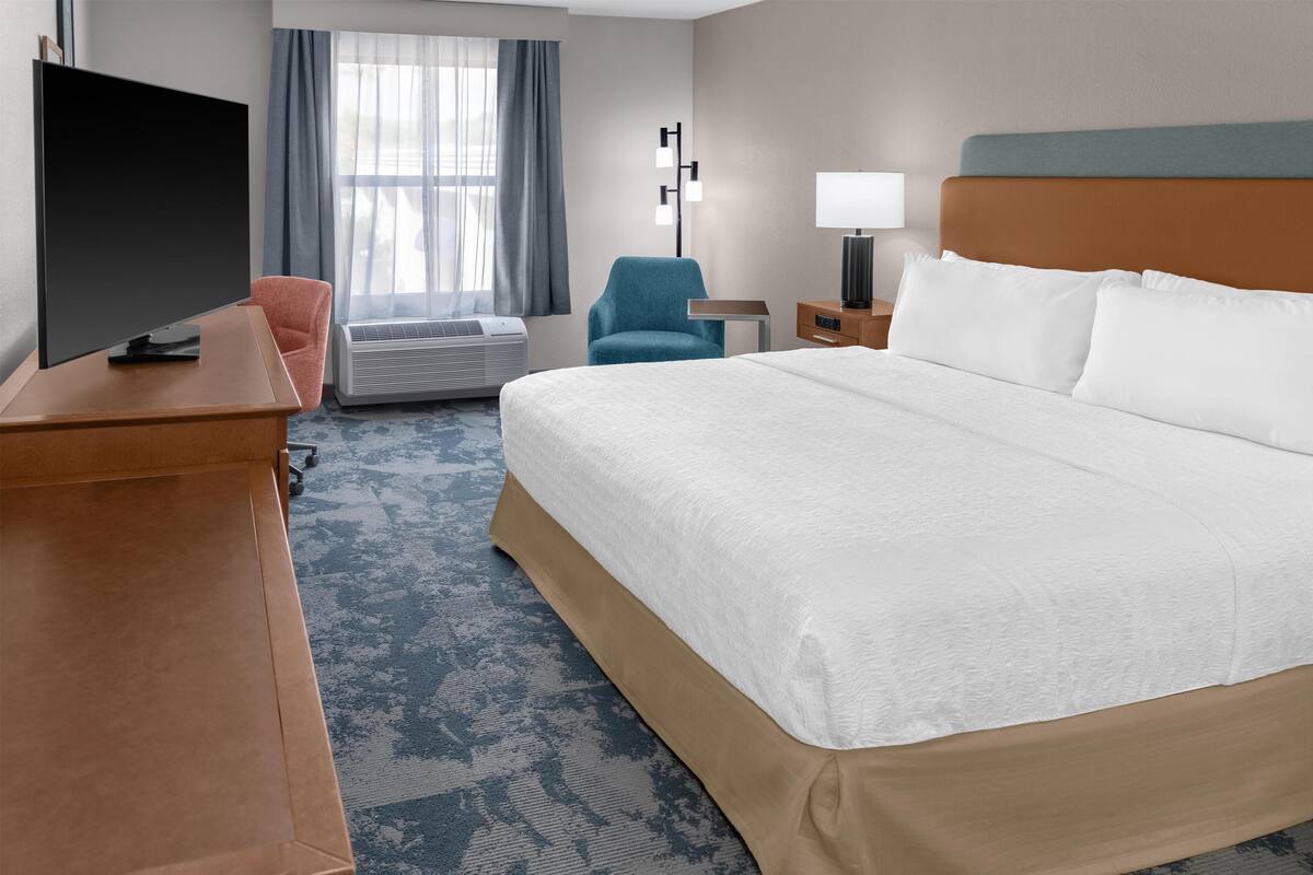 Habitación de hotel con una cama grande, un escritorio con televisor de pantalla plana, una silla, aire acondicionado y una mesita de noche con lámpara.
