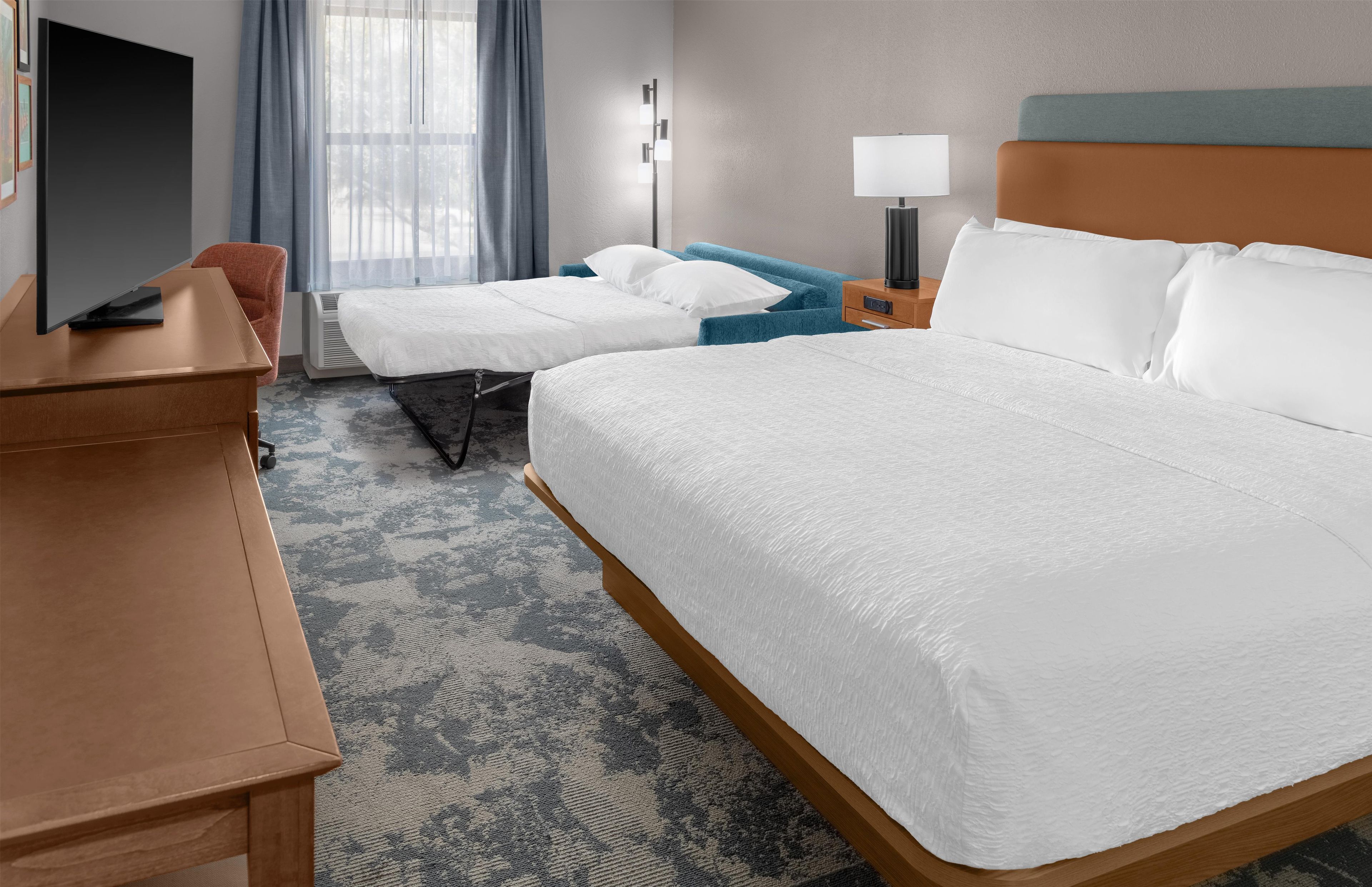 A hotel room with a large bed, a smaller bed, a desk with a flat-screen TV, a bedside table, a lamp, and a window with curtains.