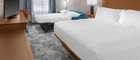 A hotel room with a large bed, a smaller bed, a desk with a flat-screen TV, a bedside table, a lamp, and a window with curtains.