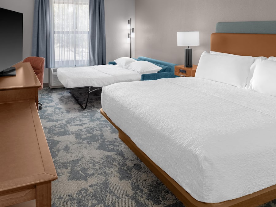 Habitación de hotel con una cama grande, otra más pequeña, un escritorio con televisión de pantalla plana, una mesita de noche, una lámpara y una ventana con cortinas.
