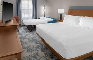 Habitación de hotel con una cama grande, otra más pequeña, un escritorio con televisión de pantalla plana, una mesita de noche, una lámpara y una ventana con cortinas.