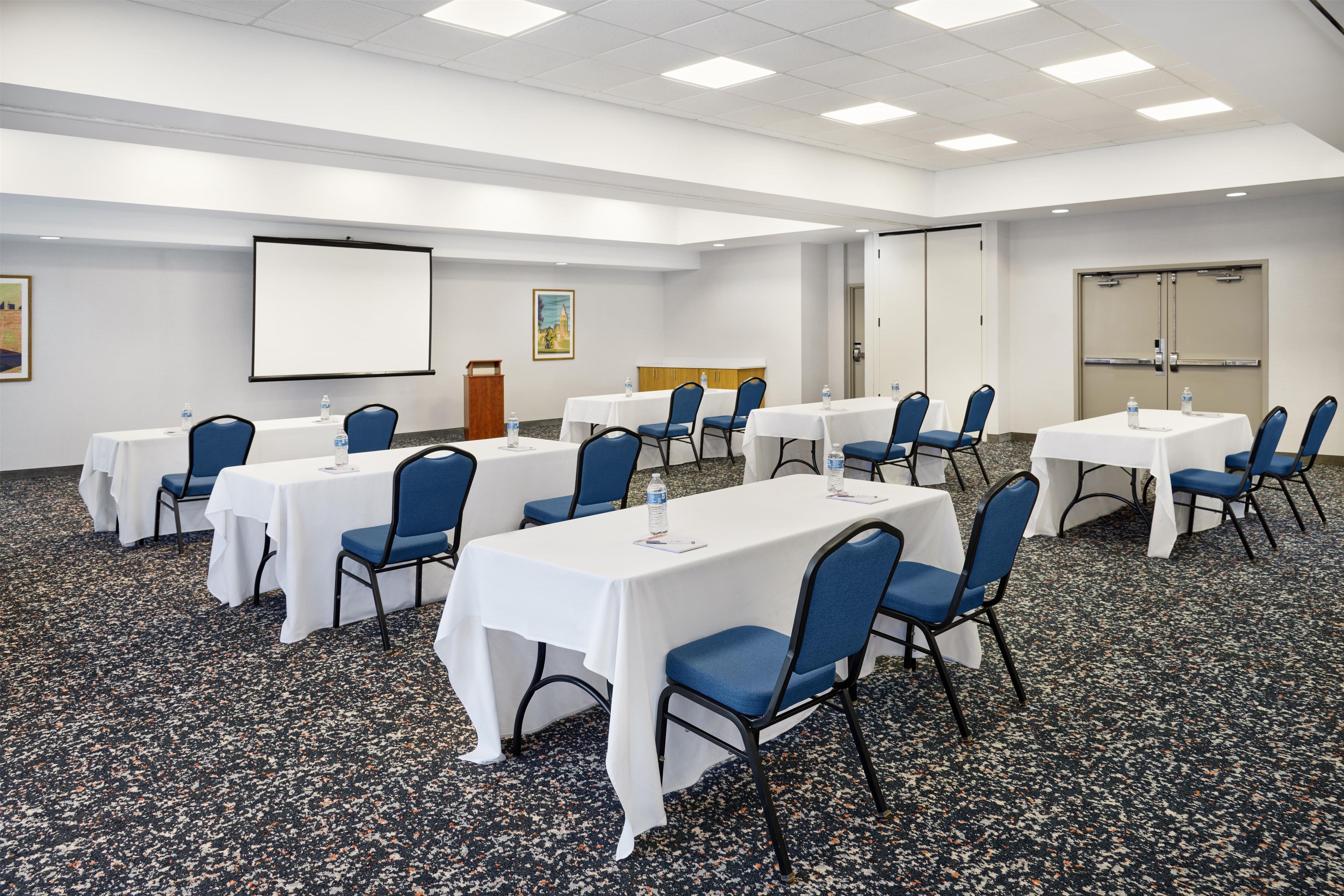 Un salón de conferencias con varias mesas y sillas, una pantalla de proyector y un dispensador de agua.