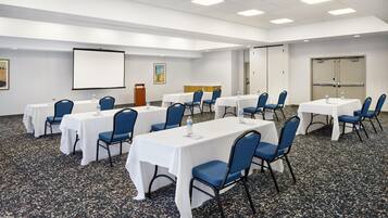 Un salón de conferencias con varias mesas y sillas, una pantalla de proyector y un dispensador de agua.
