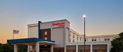 A Hampton Inn by Hilton hotel with a covered entrance, a parking lot, and an American flag.