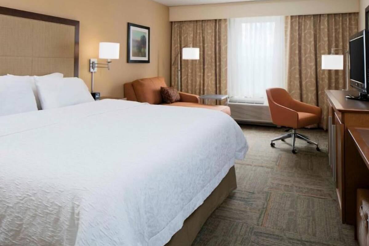 Habitación de hotel con una cama grande, un escritorio con televisor, una silla y un ventanal con cortinas.