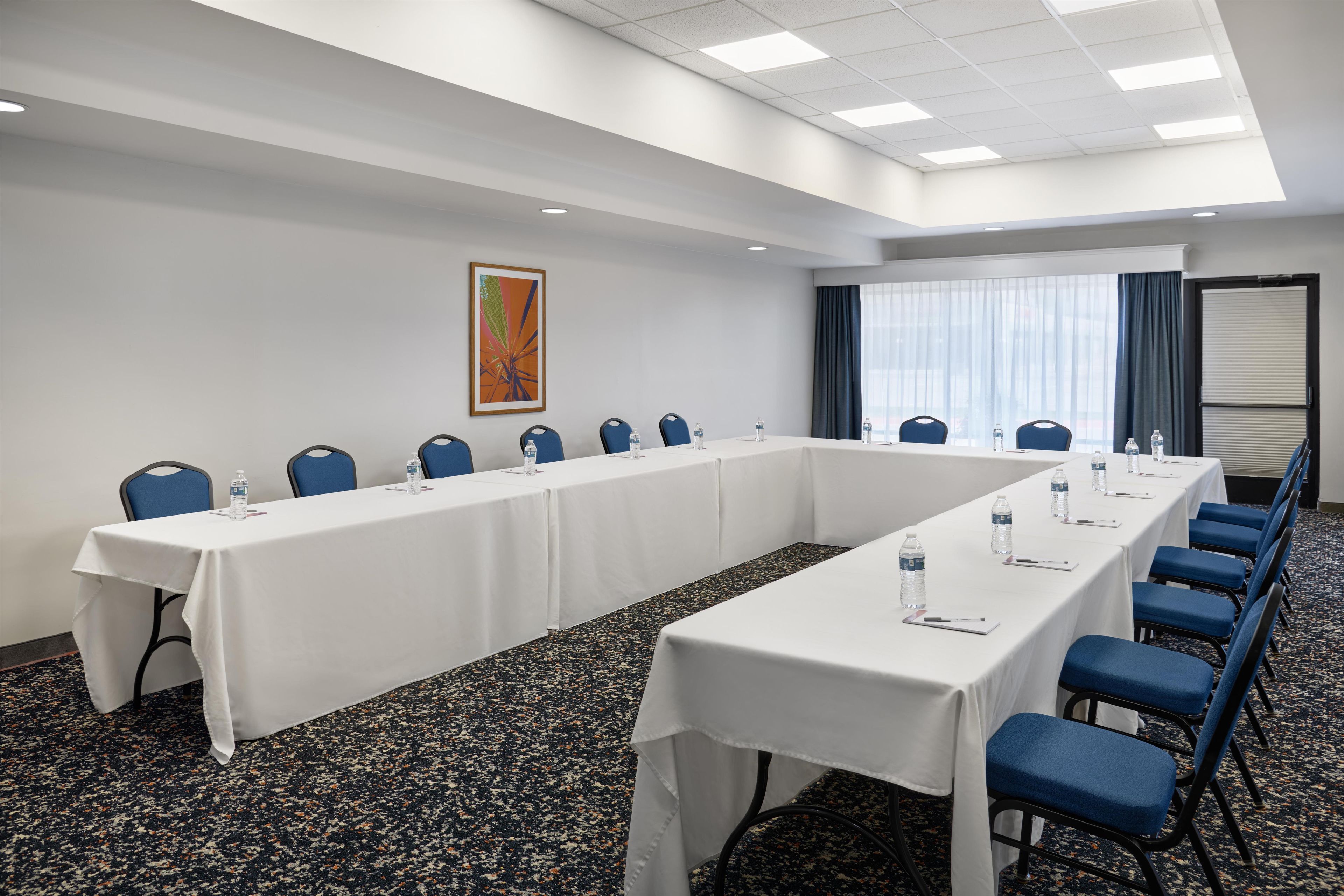 Un salón de conferencias con una larga mesa, sillas azules, botellas de agua y un cuadro en la pared.