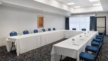 Un salón de conferencias con una larga mesa, sillas azules, botellas de agua y un cuadro en la pared.