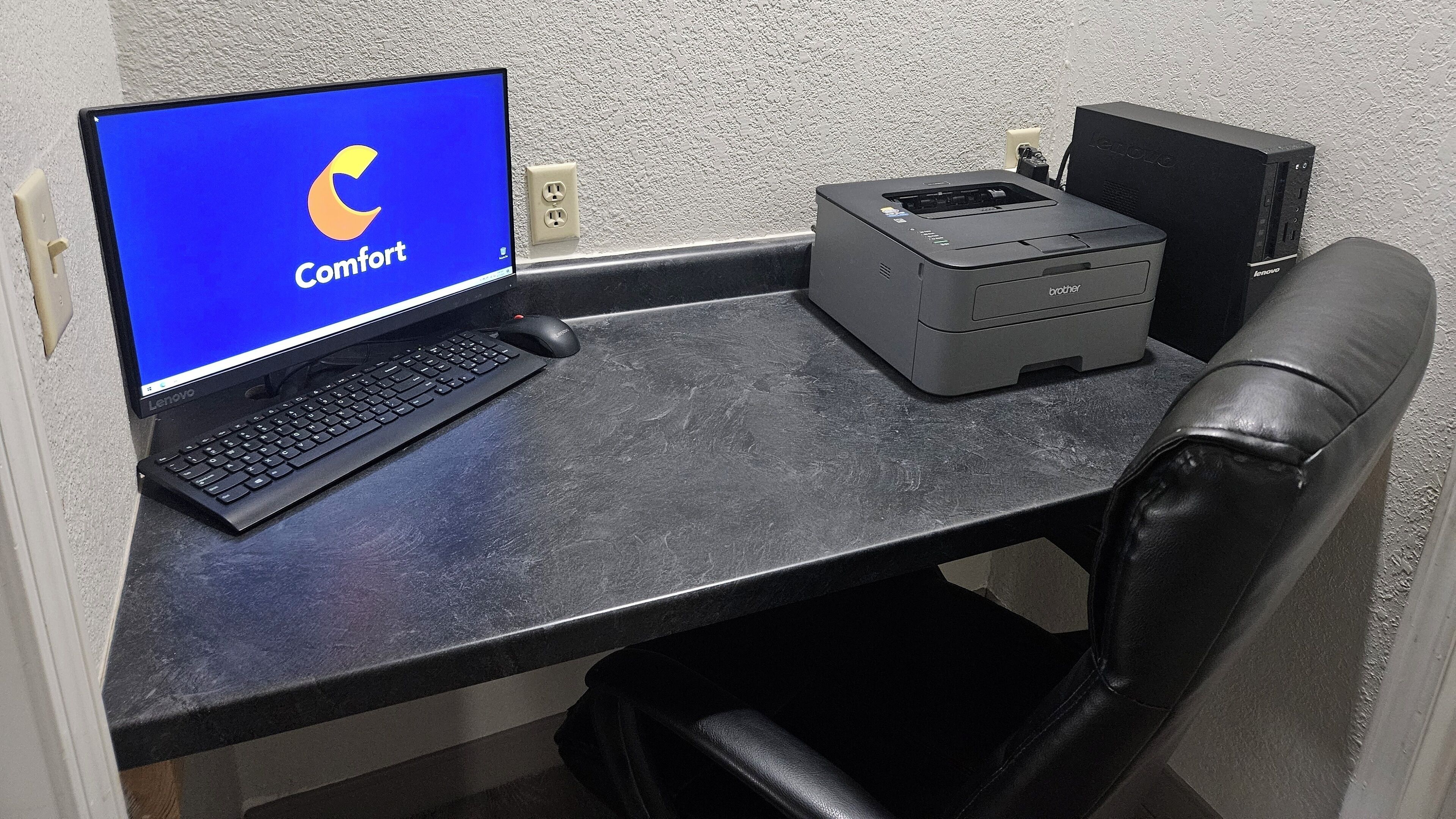 A black office chair, a black desk with a Lenovo laptop, a Brother printer, and a wall socket.