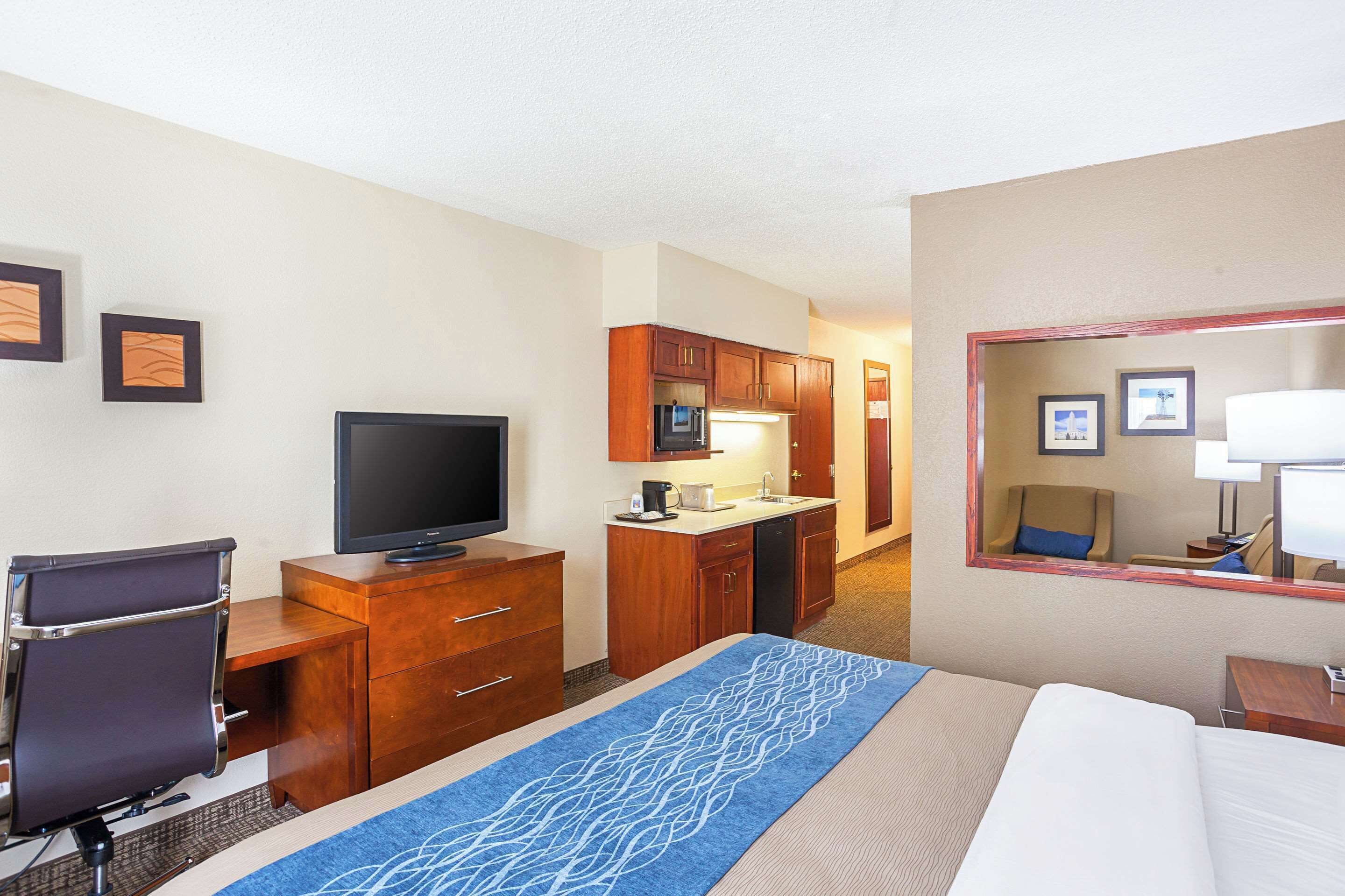 A hotel room with a bed, a desk with a TV, a kitchenette, and a mirror.