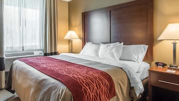 Quarto de hotel com uma cama grande, duas luminárias de cabeceira, uma cabeceira de madeira, um cobertor vermelho e uma janela com cortinas.