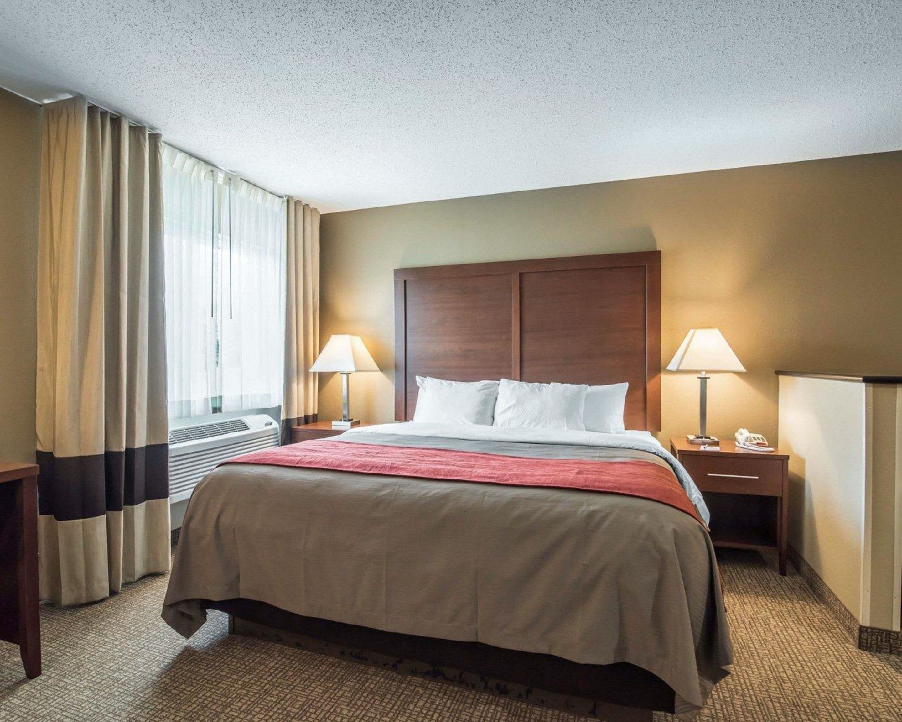 A hotel room with a large bed, two bedside tables with lamps, a wooden headboard, and a window with curtains.