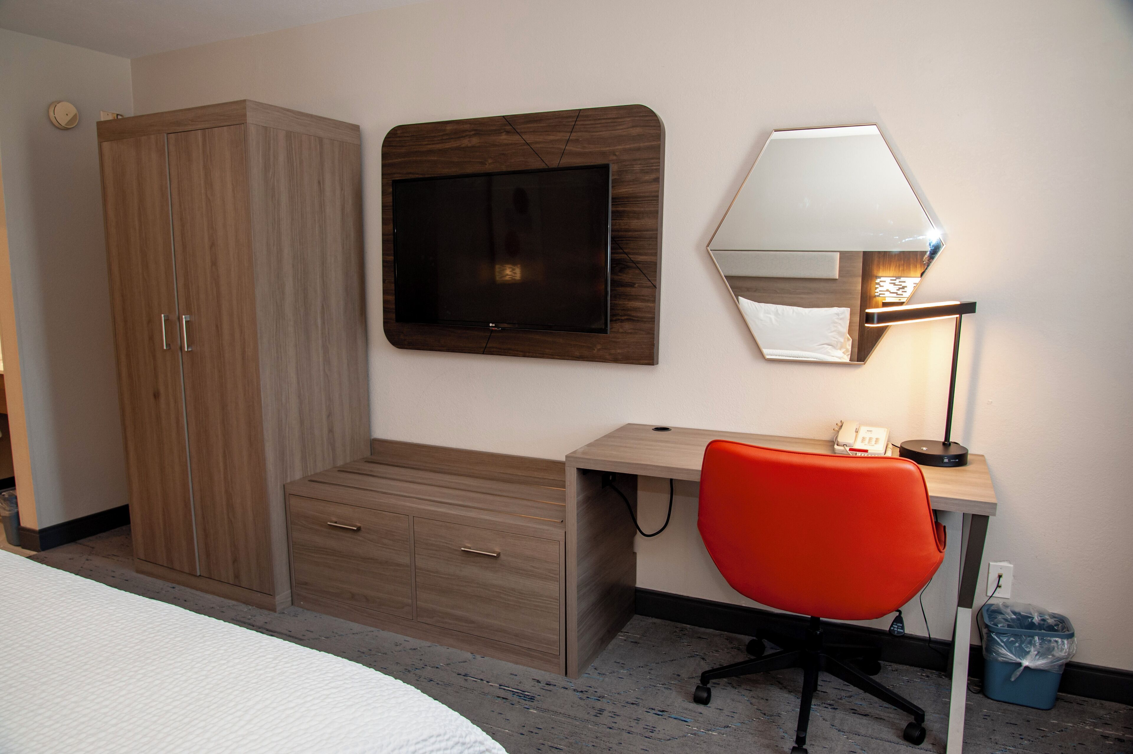 A hotel room with a bed, a desk with a chair, a TV mounted on the wall, a lamp, and a wardrobe.